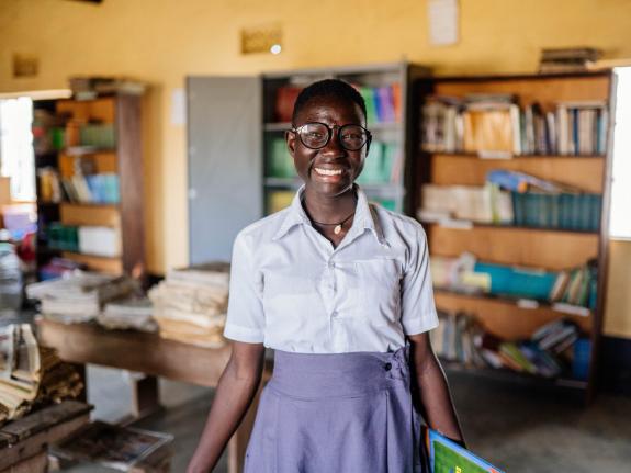 Moreen, 19, standing in her school library in Uganda surrounded by books.