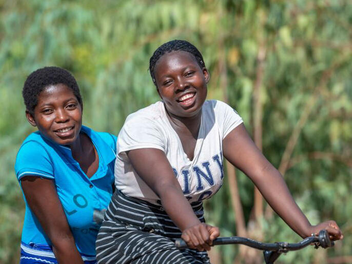  Loveness and Shakira, two young women from Malawi, riding on a bike and smiling