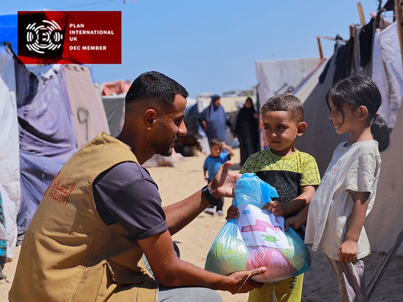 Staff member providing hot meals to 2 children with tents in background