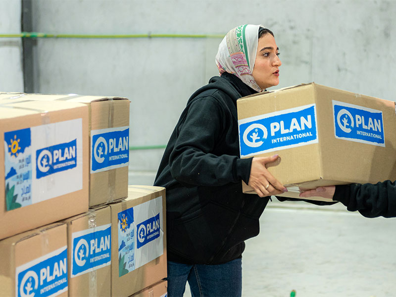 Plan International staff carrying sealed cardboard boxes with Plan International branding on the side of them. 