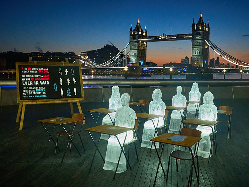 Ice sculptures of 6 girls sitting at desks with 4 desks empty with Tower Bridge in the background.