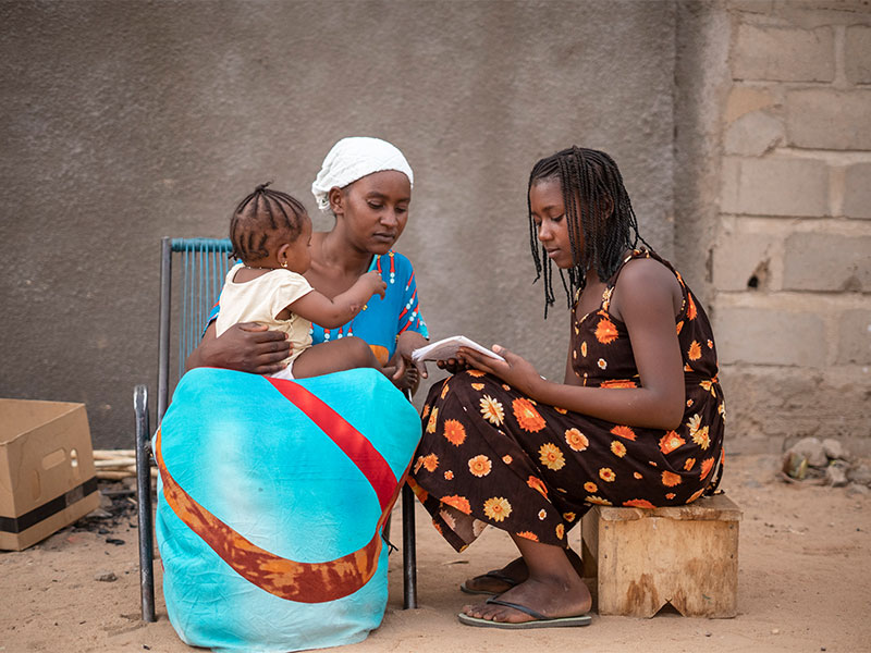 Aminata sitting outside with her mother reading from notebook. Mother has baby in lap.