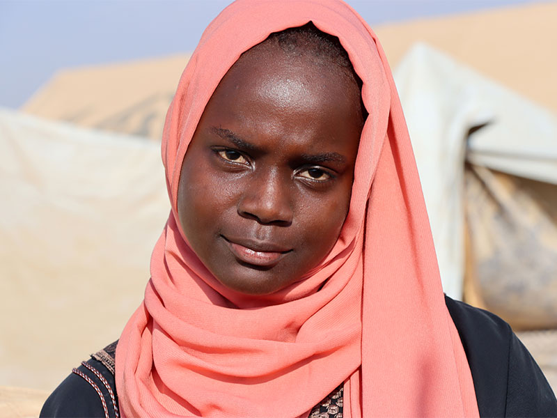Menan wearing a pink headscarf and looking at camera with tents in background.