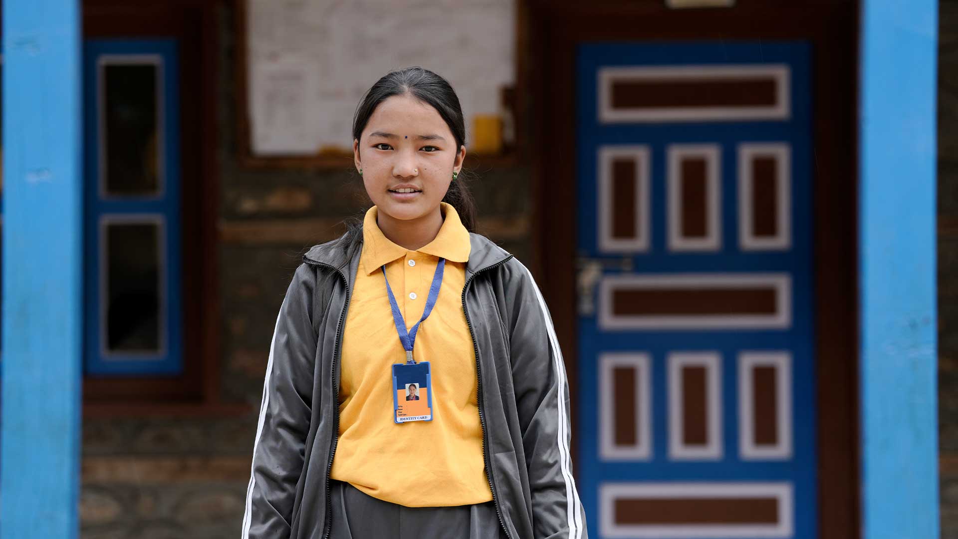 Lakshina standing in front of building and looking directly at camera wearing grey jacket and trousers with yellow polo shirt with lanyard.