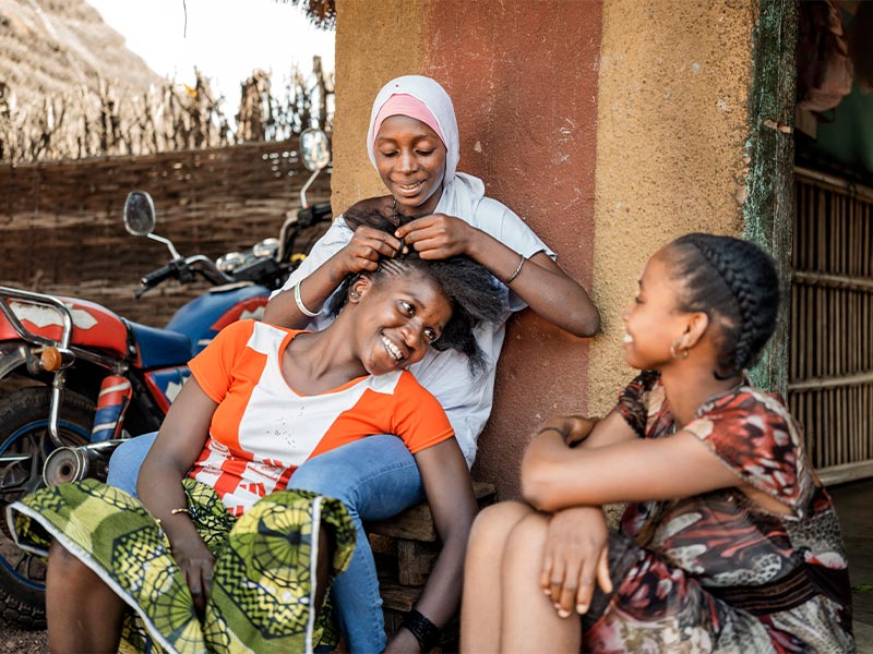Fatou braiding friend's hair while another girl is sat with them laughing and talking.