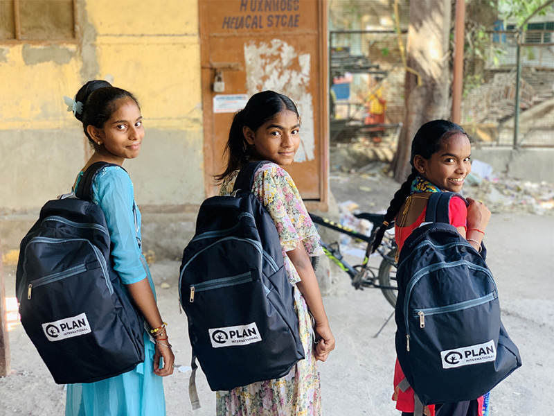 Three girls with their new education kits from Plan International. They are wearing backpacks and smiling at the camera.