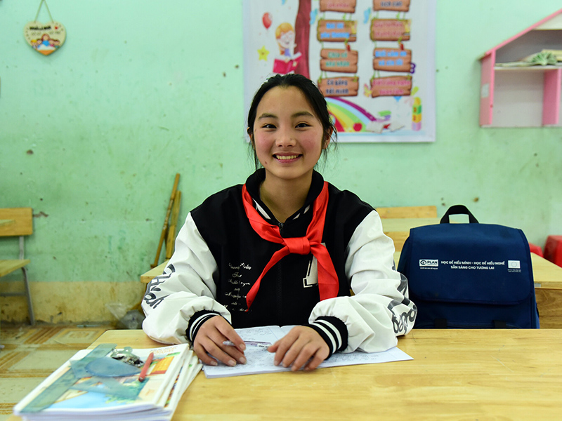 Sào, from Vietnam, sits in a classroom. She is working on a workbook, with a school bag beside her.