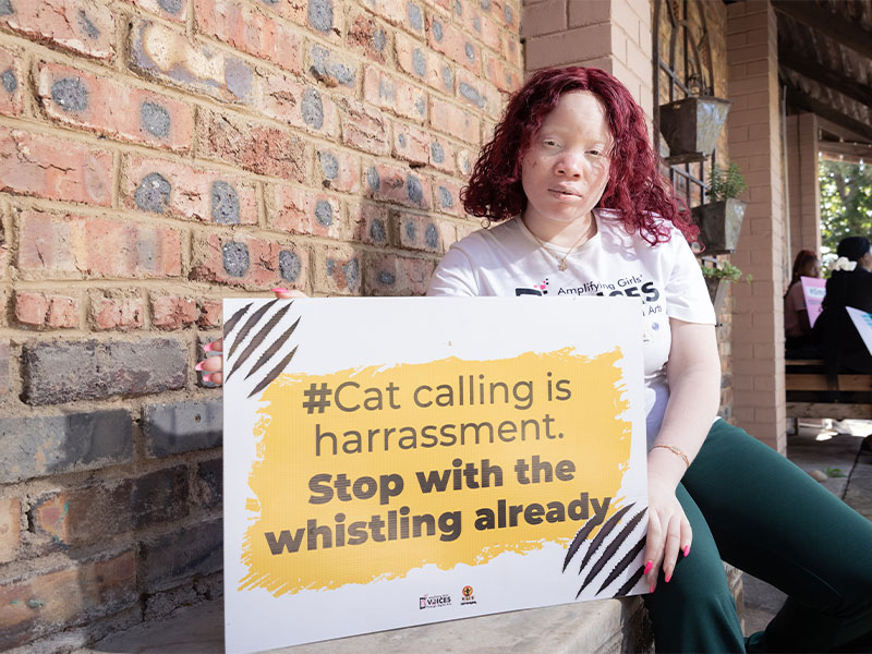 Sisasenkosi sat outside holding a sign that says "Cat calling is harassment. Stop with the whistling already."