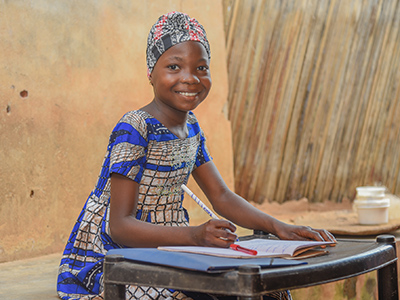 Anne, 12, writes in her school workbook, sitting at a desk. She is wearing a blue dress.