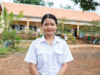 Phgnon, 16, stands in front of her school. She is wearing a white school shirt.