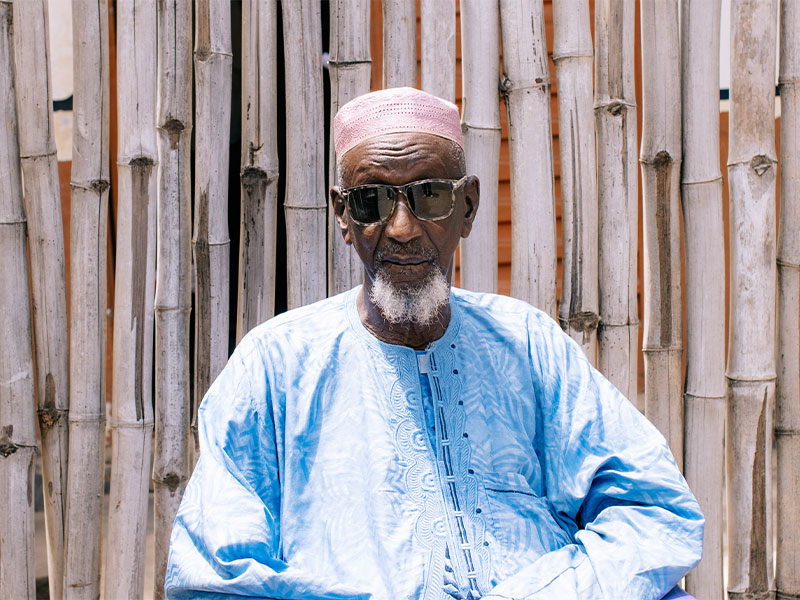 Imam Sheik Ibrahim sat outside with wooden wall behind him
