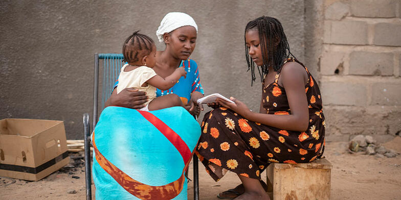 Young girl sitting with mother and sibling, reading from book. 