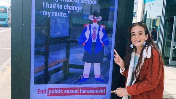 Jess stands next to one of the Crime Not Compliment adverts, calling for public sexual harassment to be made a crime