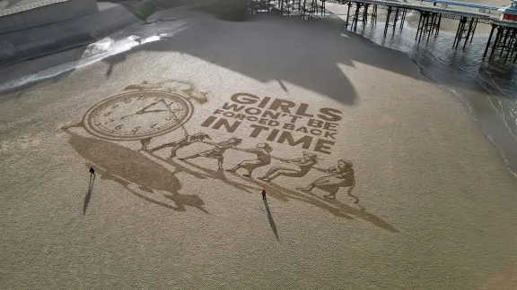 Sand art showing girls pulling on a rope attached to a clock with words 'girls won't be forced back in time'