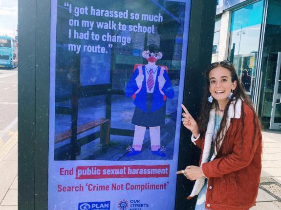 Jess stands next to one of the Crime Not Compliment adverts, calling for public sexual harassment to be made a crime