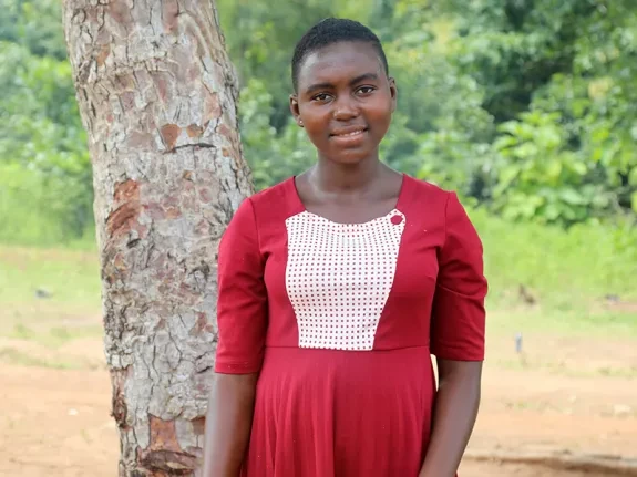 Dorcas, 16, from Togo, stands next to a tree. She is wearing a red dress and smiling at the camera.