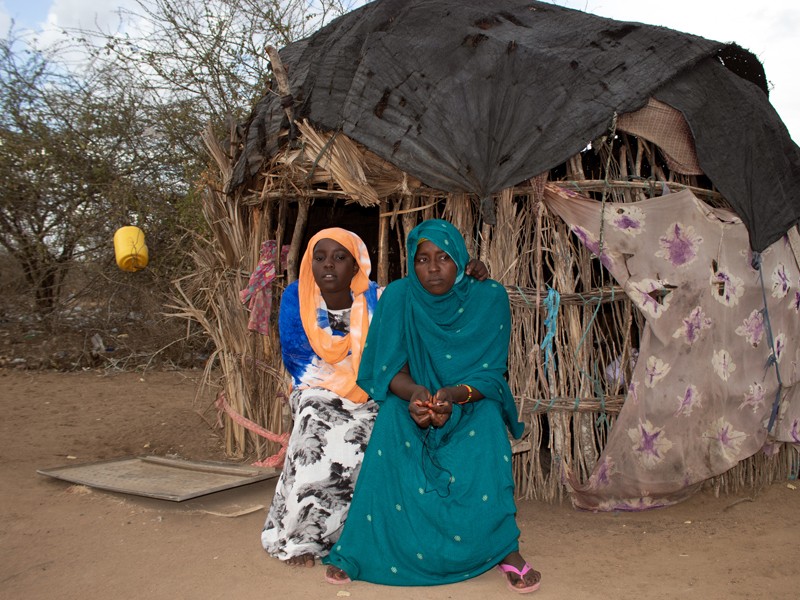 Najma and her mother Lulu are sitting in front of a brown hut that is their home