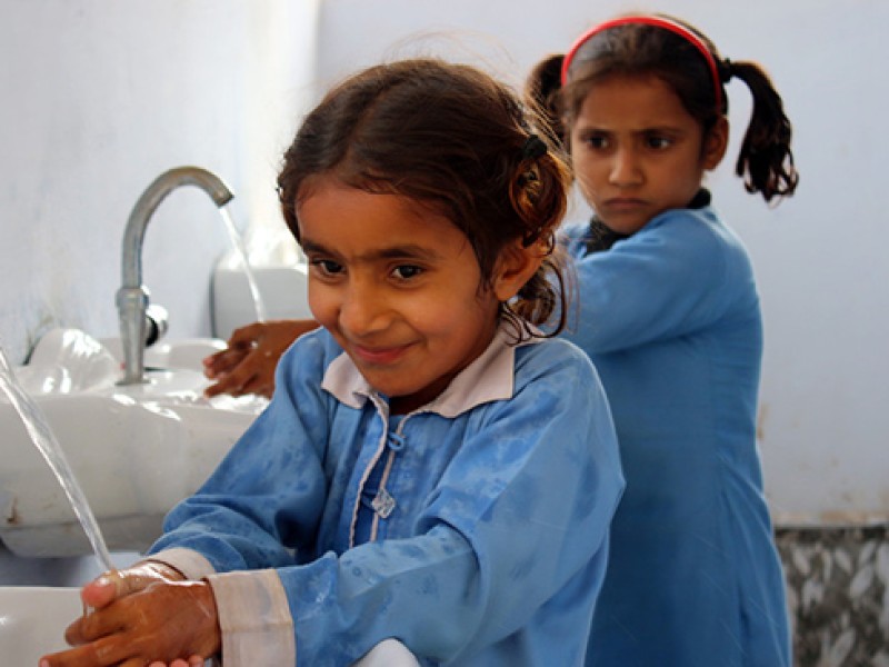 Two young girls in school uniforms wash their hands in basins with taps