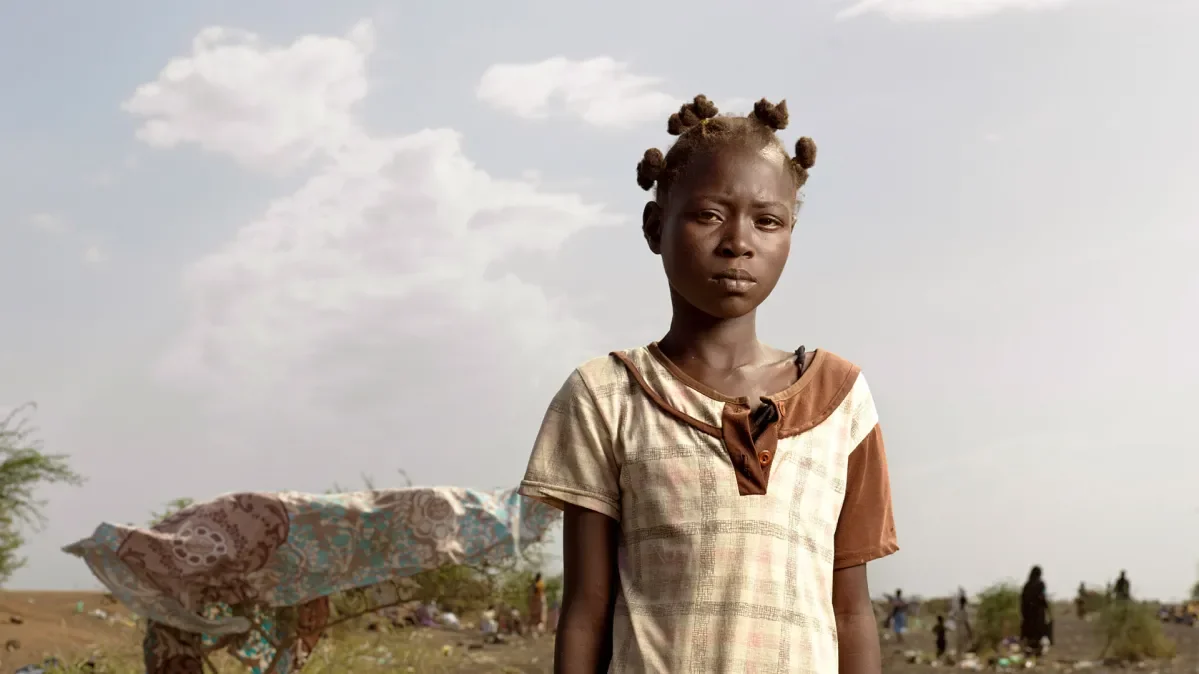 Jacklyn standing outside looking at camera with group of adults and children sat behind her. 