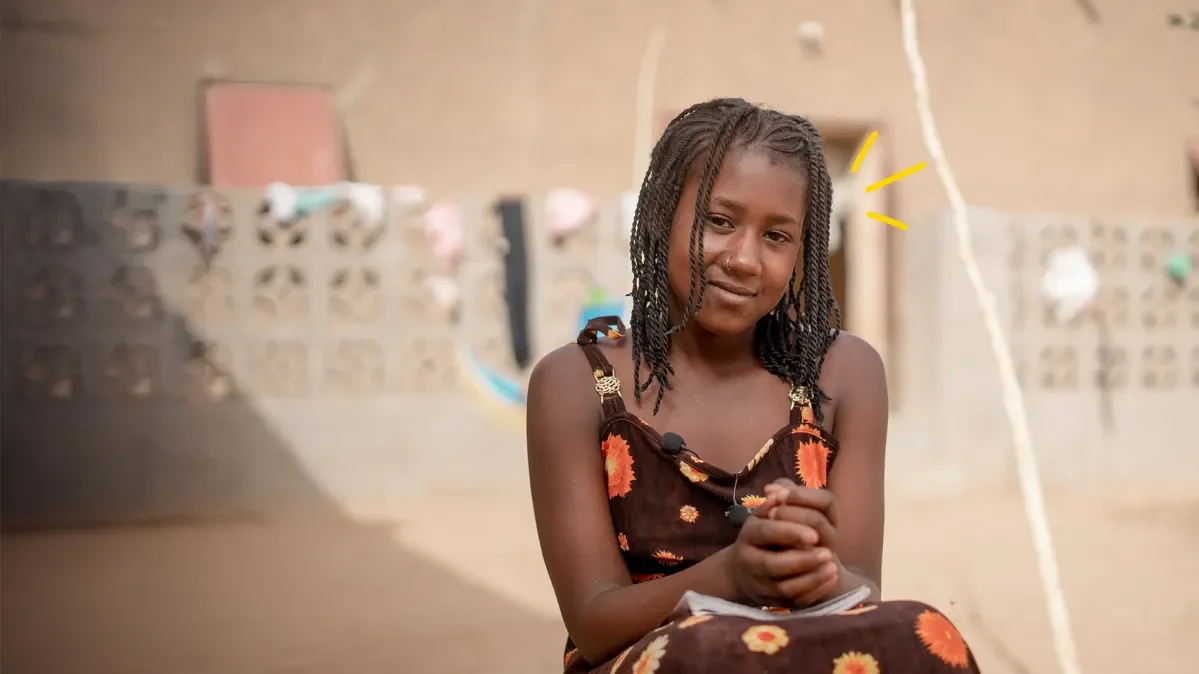 Young girl sitting outside with book on lap and hands clasped together on top of it smiling at camera