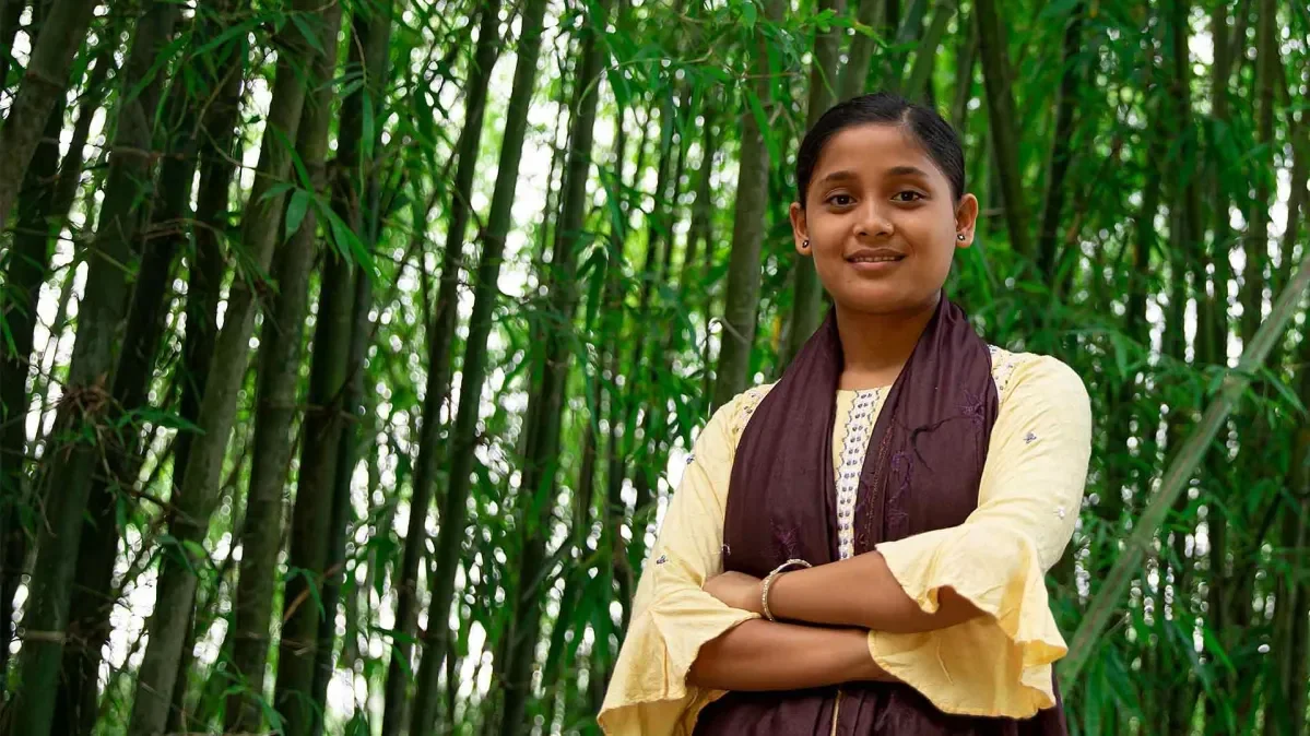 Antora, 16, standing against a background of bamboo trees in her home district of Rangpur in Bangladesh.