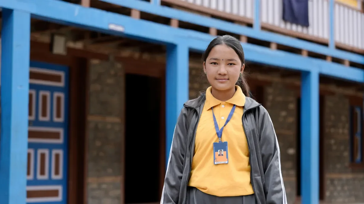 Lakshina standing in front of building and looking directly at camera wearing grey jacket and trousers with yellow polo shirt with lanyard.