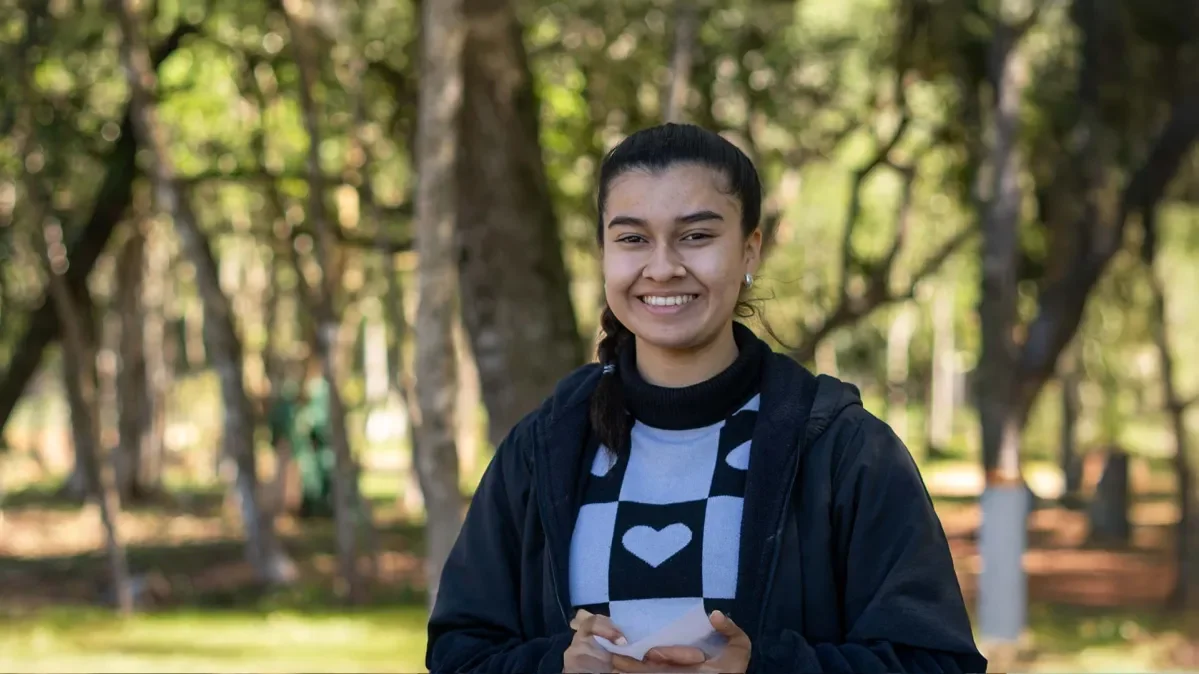 Rebecca standing outside smiling at camera with out of focus trees behind her