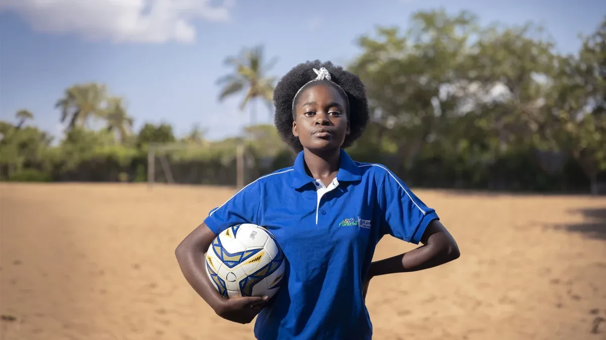 Eunice, from Mozambique, stands on a football pitch. She is wearing a blue shirt and holding a football.
