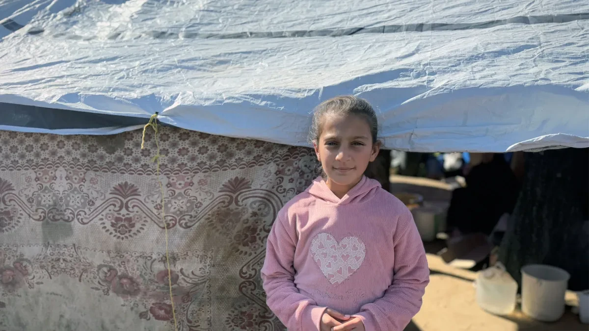 Elin, 11, in Northern Gaza, stands in front of a tent. She is wearing a pink hoodie.