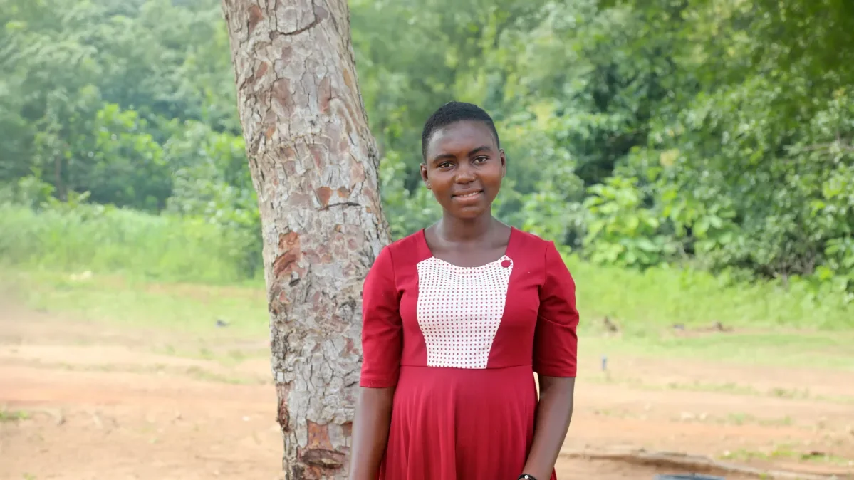 Dorcas, 16, from Togo, stands next to a tree. She is wearing a red dress and smiling at the camera.