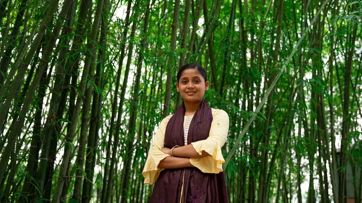 Antora, 16, standing against a background of bamboo trees in her home district of Rangpur in Bangladesh.