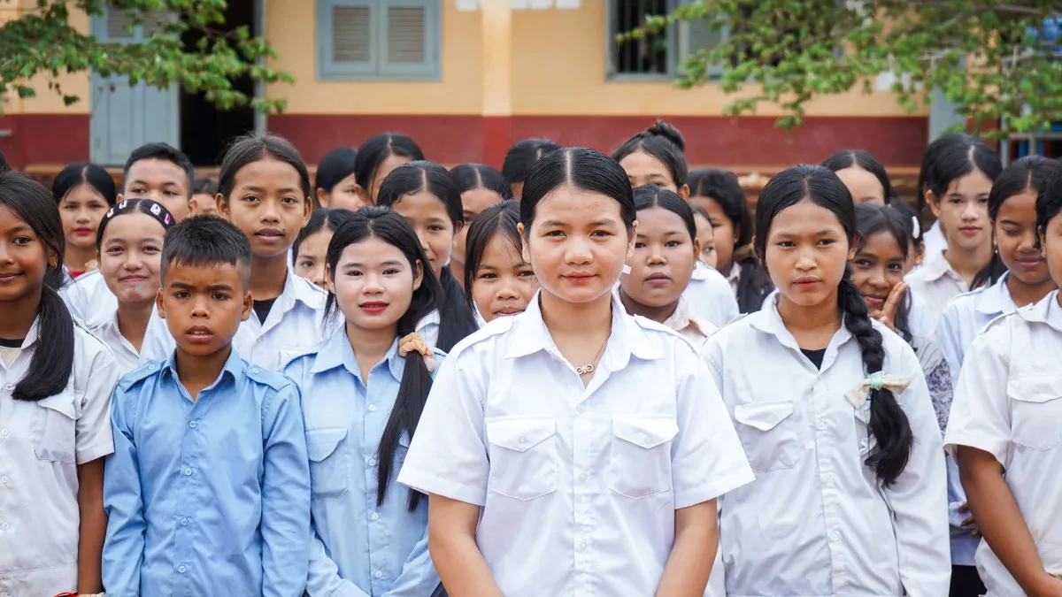 Phgnon, 16, stands with her classmates in front of their school. They are wearing white school shirts.