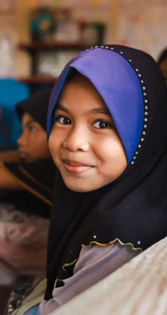 Young girl in classroom smiling at camera with other students blurred in background.