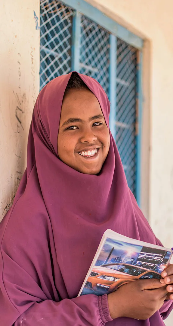 Hamda, standing in front of wall holding book and smiling at camera