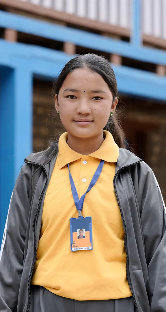 Lakshina standing in front of building and looking directly at camera wearing grey jacket and trousers with yellow polo shirt with lanyard.