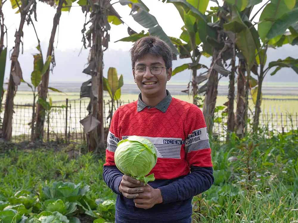 Shimul, 16, standing in his family's crop garden holding a cabbage