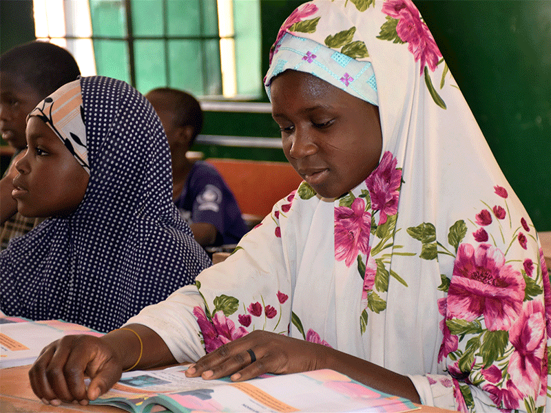 Khadija sat at desk reading book on her table with other pupils in the background. 