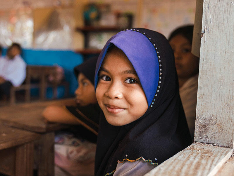 Young girl in classroom smiling at camera with other students blurred in background.