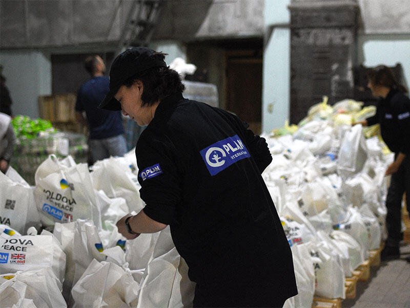 Volunteers wearing Plan International branded jackets moving/ sorting parcels.