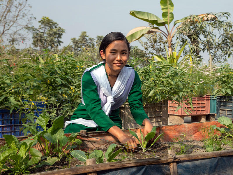 Young girl sat down tending to vegetable garden looking at camera.