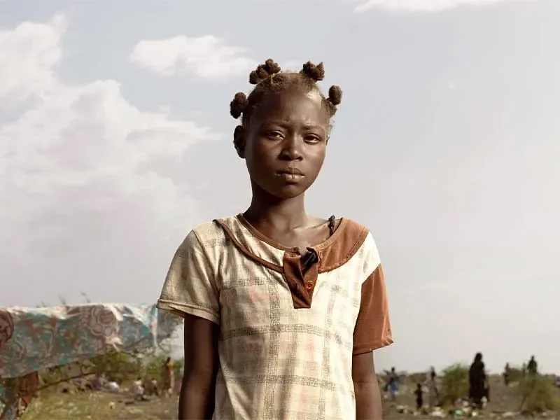 Jacklyn standing outside looking at camera with group of adults and children sat behind her. 