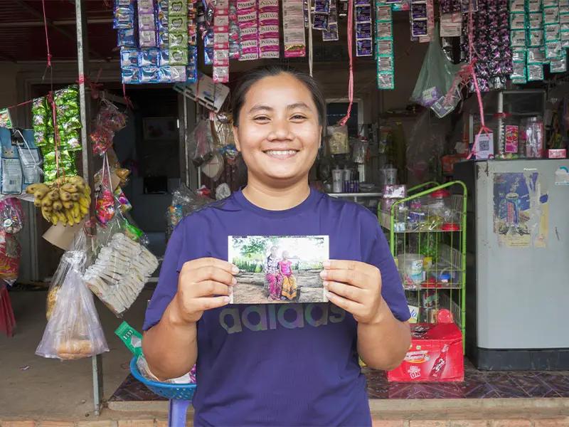 Ousa smiling at camera and holding a photo of herself as a young girl towards viewer.