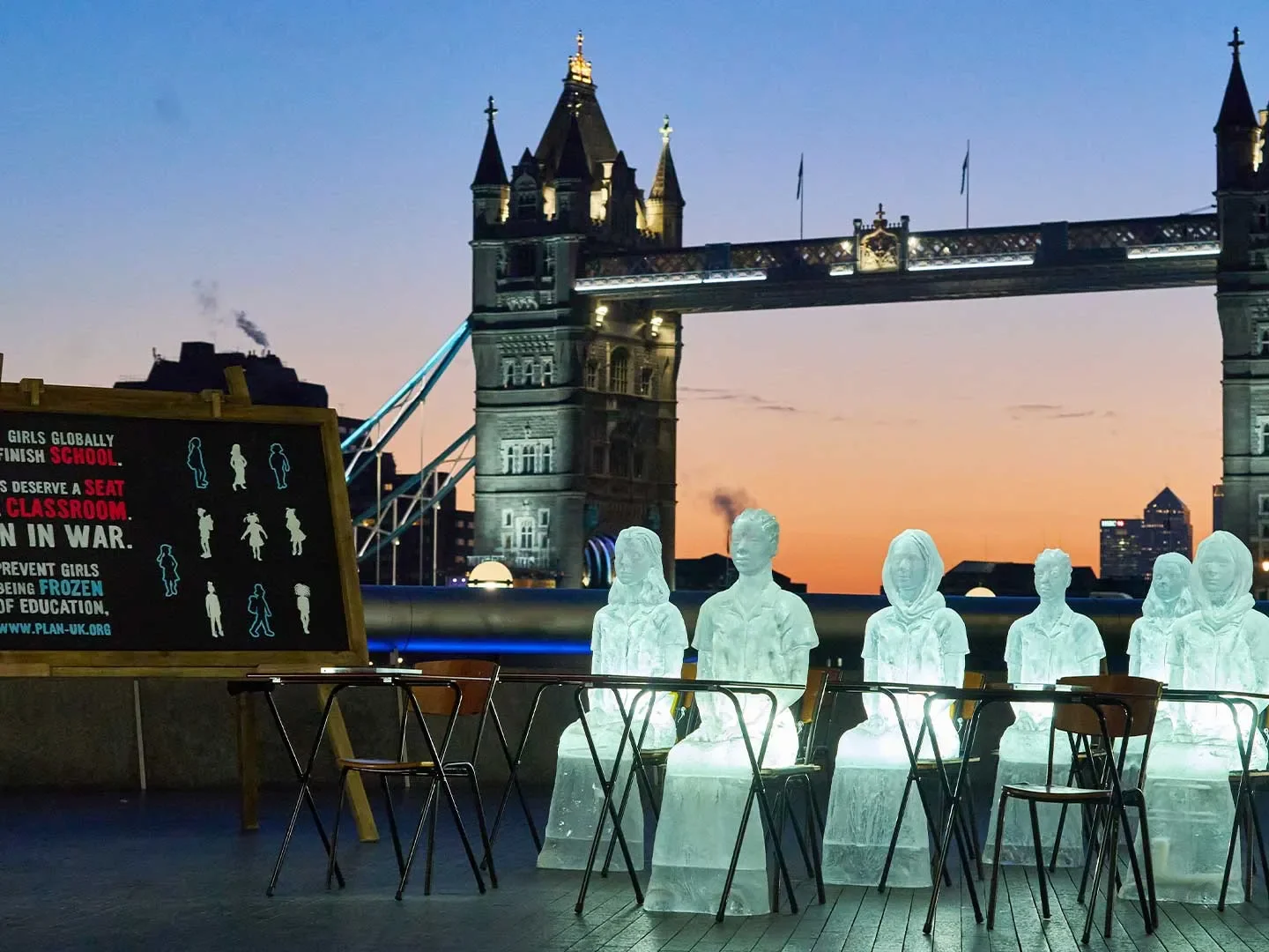 Ice sculptures of 6 girls sitting at desks with 4 desks empty with Tower Bridge in the background.