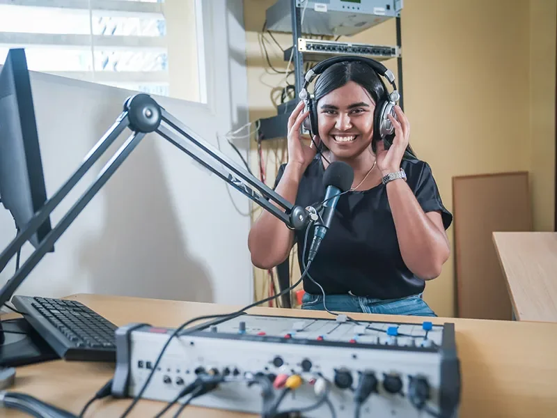 Edith stands in front of radio equipment. She is wearing headphones and smiling. 