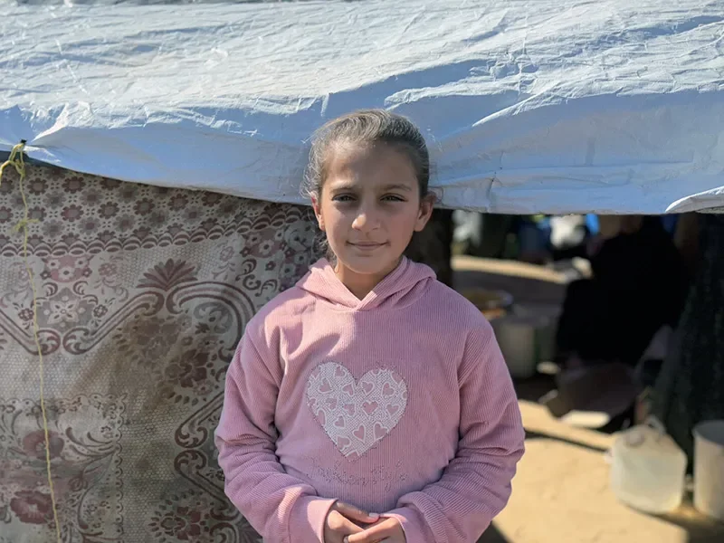 Elin, 11, in Northern Gaza, stands in front of a tent. She is wearing a pink hoodie.