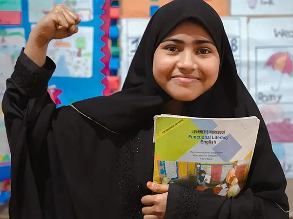 Ayesha, aged 14, stands in a classroom and flexes her arm. She is holding an English literacy textbook.