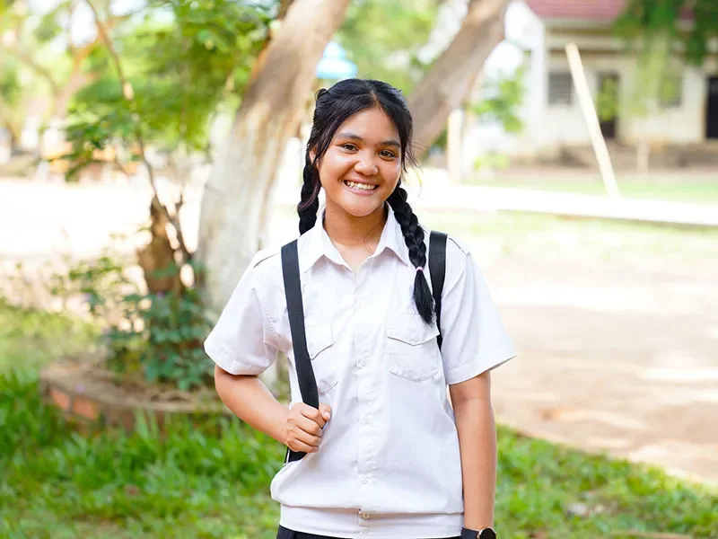 Sopheak, 15, from Cambodia, stands in front of a tree. She is wearing a school rucksack on her back.