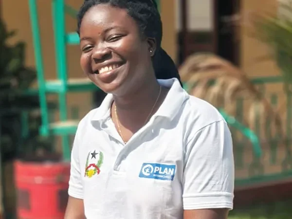 Aissatu, aged 21, wears a white shirt with a Plan International logo. She is smiling at the camera.