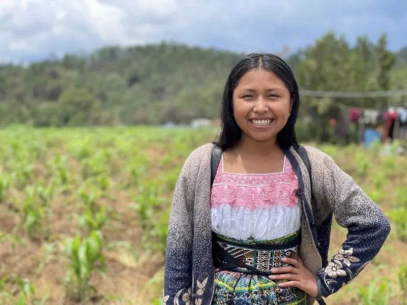 Kimberly, from Guatemala, stands in a field. She has a hand on her hip and smiles at the camera.