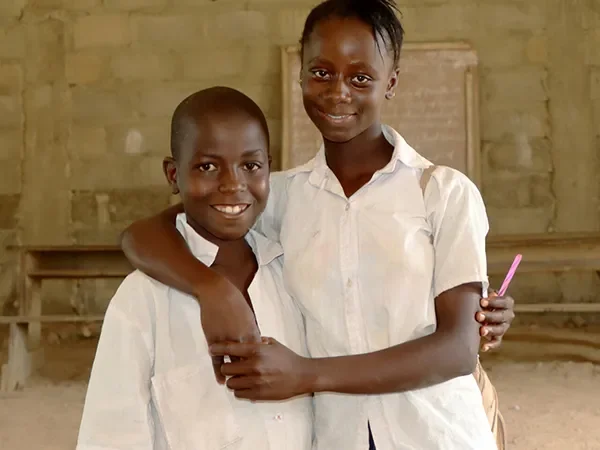 Ibrahim and Marita, from Liberia, stand side by side in a school. They have their arms around each other and smile at the camera.